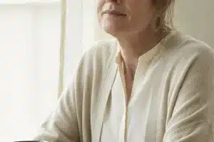 Frau Anfang 50 mit zurückgebundenem aschblondem Haar und hellem Cardigan sitzt mit dunklem Becher am Fenster