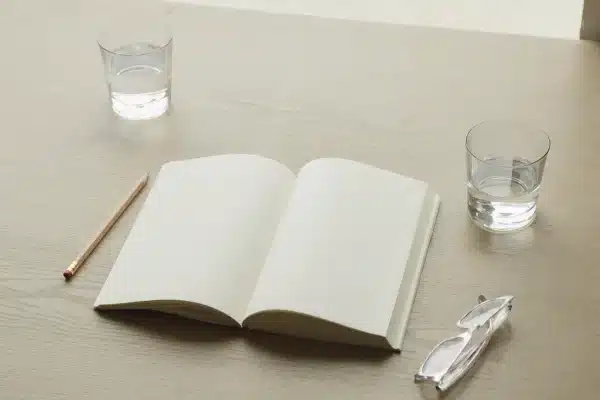 Aufgeschlagenes leeres Notizbuch, daneben Bleistift, Lesebrille und zwei Wassergläser auf einem hellen Eichentisch im Tageslicht