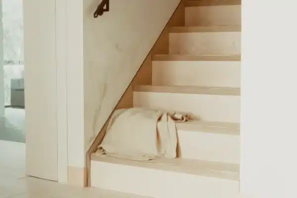 Helle Holztreppe mit dunklem Metallhandlauf, auf der untersten Stufe ein locker gefaltetes Leinenplaid, links Durchblick zum Fenster