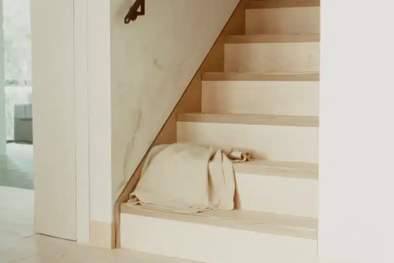 Helle Holztreppe mit dunklem Metallhandlauf, auf der untersten Stufe ein locker gefaltetes Leinenplaid, links Durchblick zum Fenster