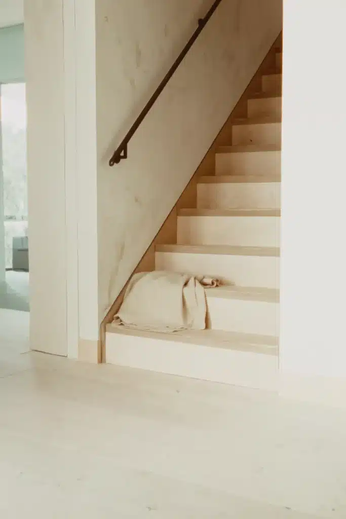 Helle Holztreppe mit dunklem Metallhandlauf, auf der untersten Stufe ein locker gefaltetes Leinenplaid, links Durchblick zum Fenster