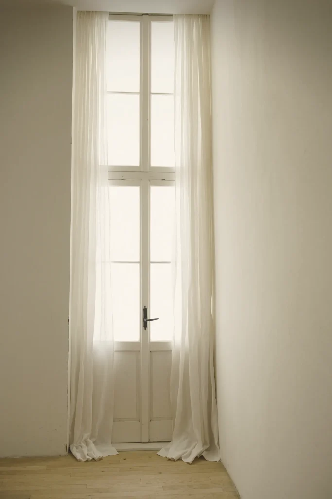 Hohe Fenstertür mit halbtransparenten weißen Vorhängen, die bis auf den hellen Holzboden fallen, im Tageslicht