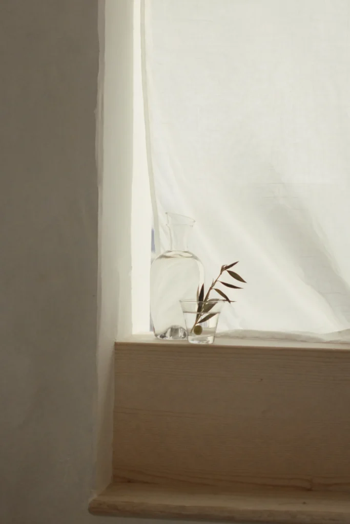 Glaskaraffe und Wasserglas mit Olivenzweig auf einer hellen Holzfensterbank vor einem Leinenvorhang im Gegenlicht