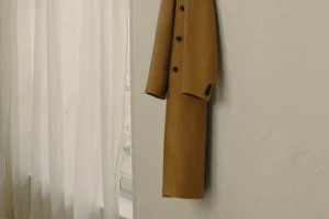 Camelfarbener Wollmantel an einem dunklen Wandhaken in einem hellen Raum neben einem Fenster mit weißem Vorhang