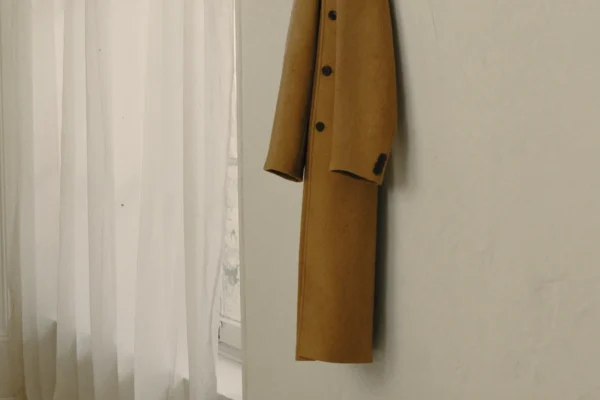 Camelfarbener Wollmantel an einem dunklen Wandhaken in einem hellen Raum neben einem Fenster mit weißem Vorhang