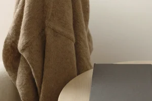 Hellbraune Strickjacke über der Lehne eines hellen Polstersessels, daneben runder Holztisch mit grauem Buch