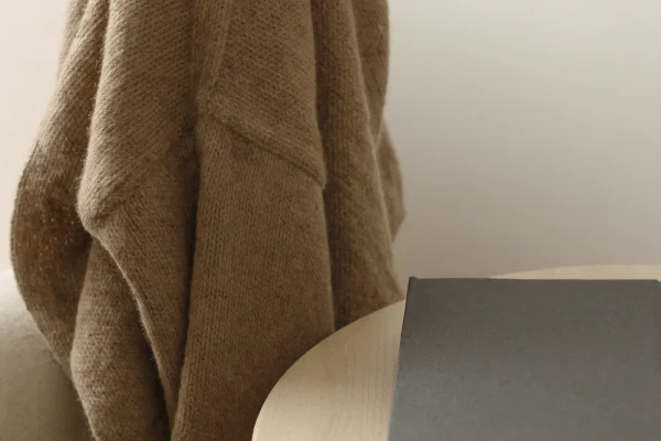 Hellbraune Strickjacke über der Lehne eines hellen Polstersessels, daneben runder Holztisch mit grauem Buch