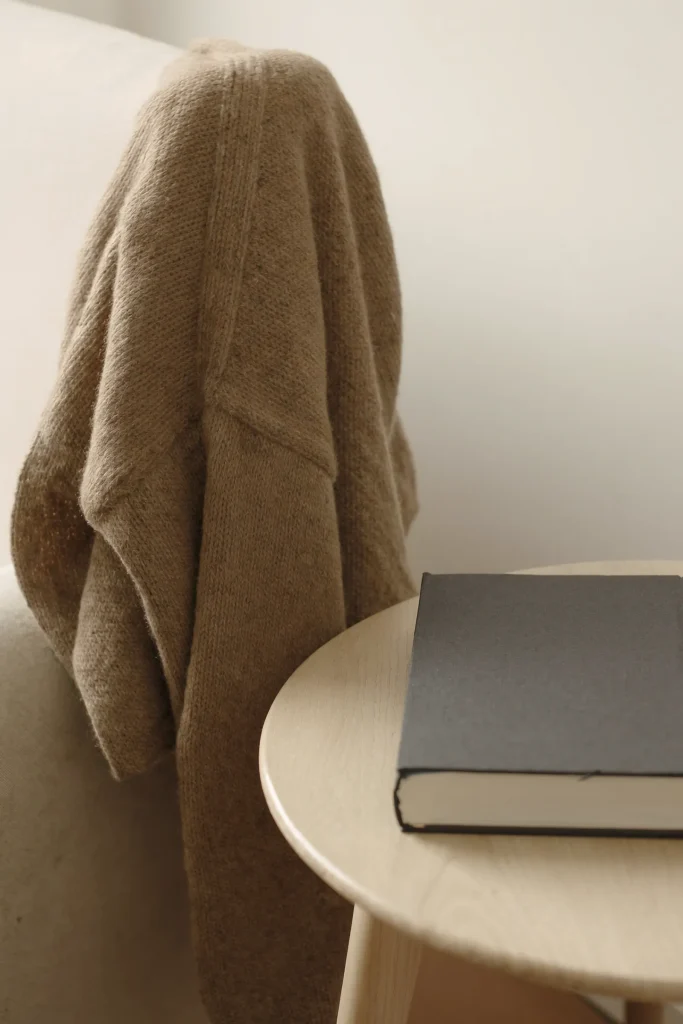 Hellbraune Strickjacke über der Lehne eines hellen Polstersessels, daneben runder Holztisch mit grauem Buch