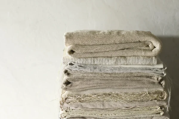 Stapel gefalteter Leinen- und Wollstoffe in Creme, Sand und Grau auf einem hellen Holzbrett vor verputzter Wand