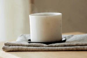 Dampfende weiße Tasse auf schwarzem Untersetzer, darunter ein gefaltetes Leinentuch auf hellem Holztisch im Tageslicht
