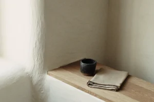 Dunkler Keramikbecher und gefaltete beige Leinenserviette auf einem Holzbrett in einer weiß verputzten Wandnische im Tageslicht
